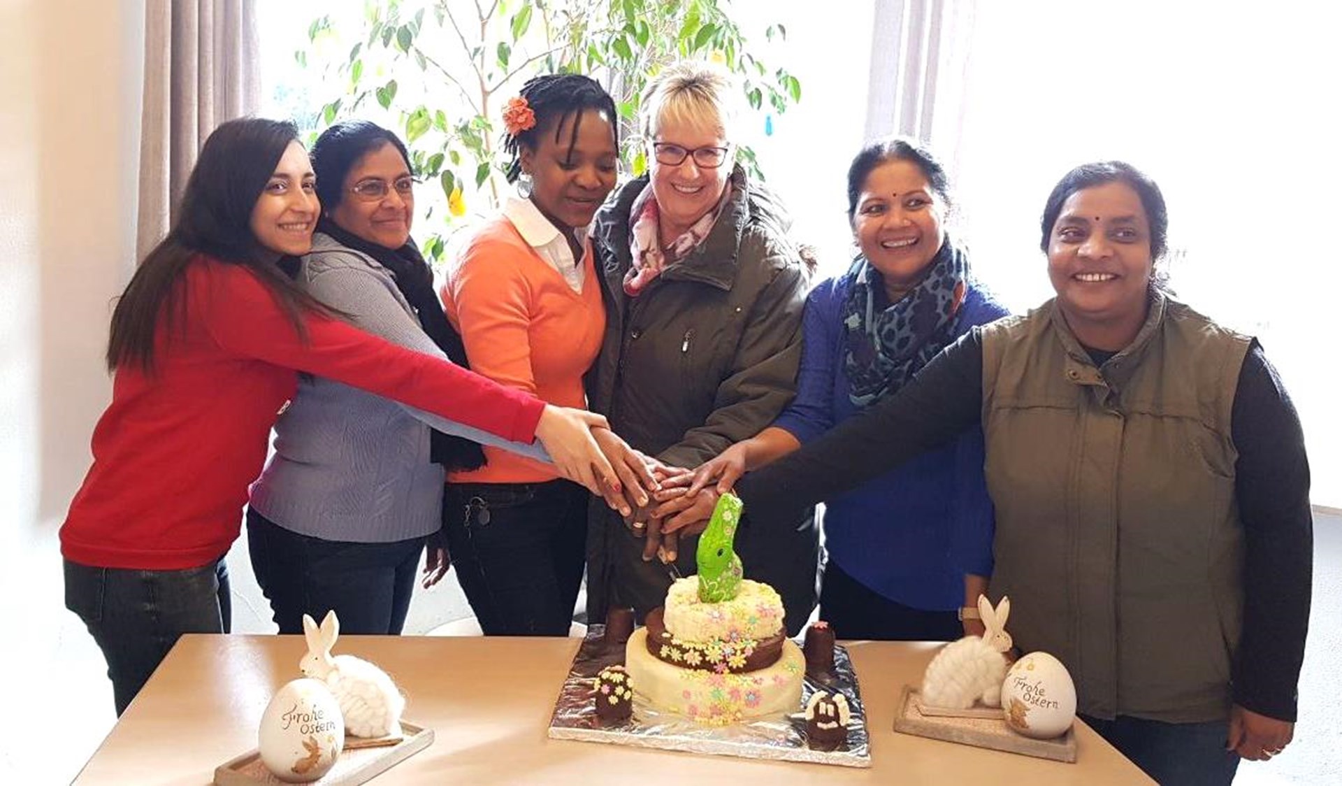 Sechs Frauen unterschiedlicher Kulturen stehen nebeneinander und reichen sich die Hände. Davor steht auf einem Tisch eine verzierte Torte mit einem Schokoladen-Osterhasen.