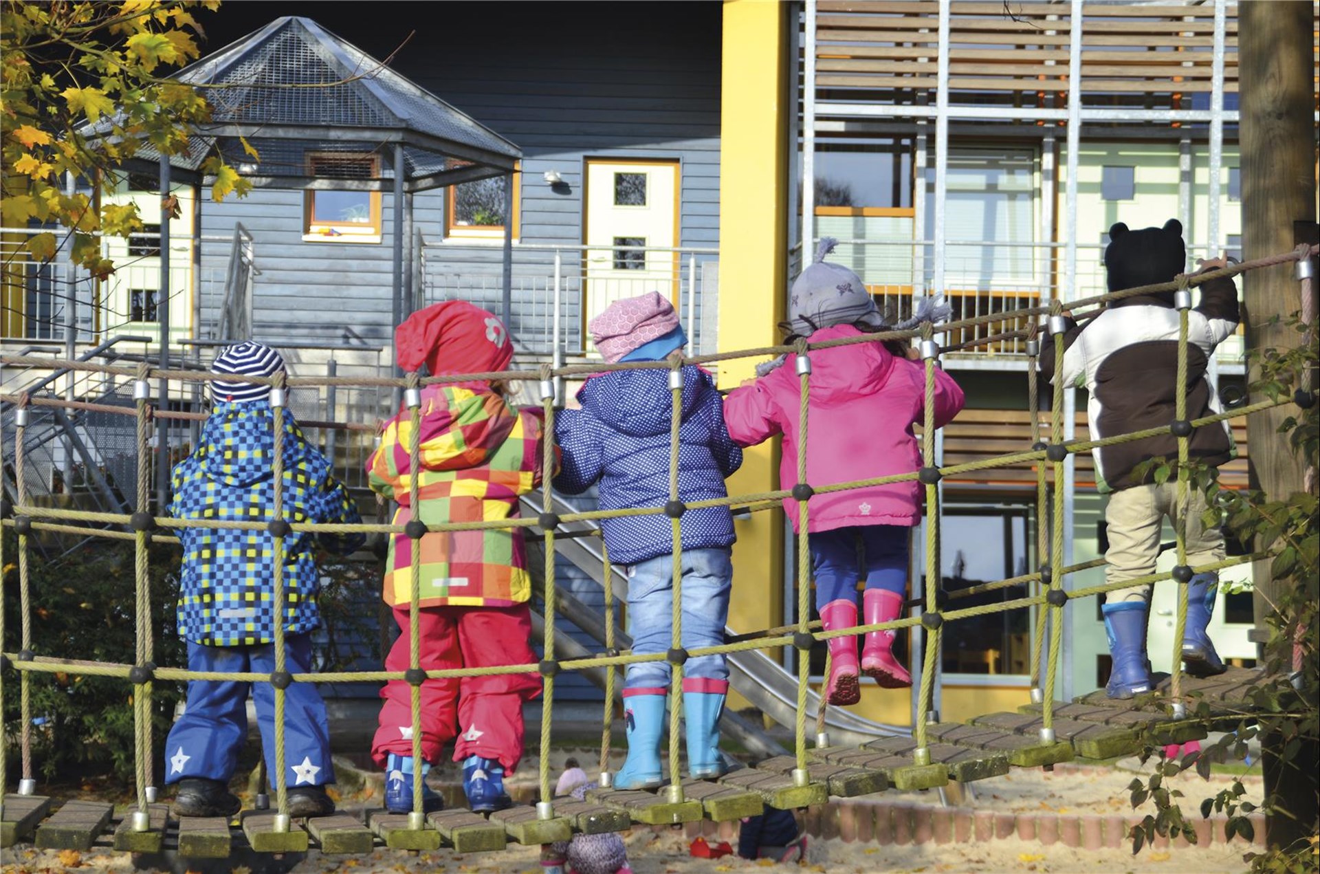 Kinder stehen auf einer Hängebrücke auf dem Spielplatz, zu sehen von hinten