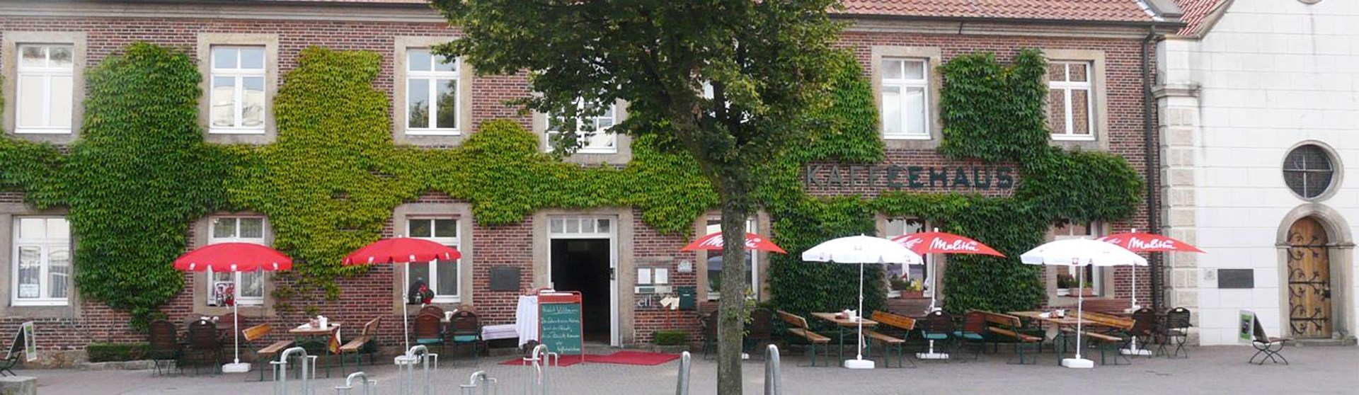 Backsteingebäude mit vielen Fenstern und einer Terrasse davor mit Tischen, Stühlen und Sonnenschirmen.