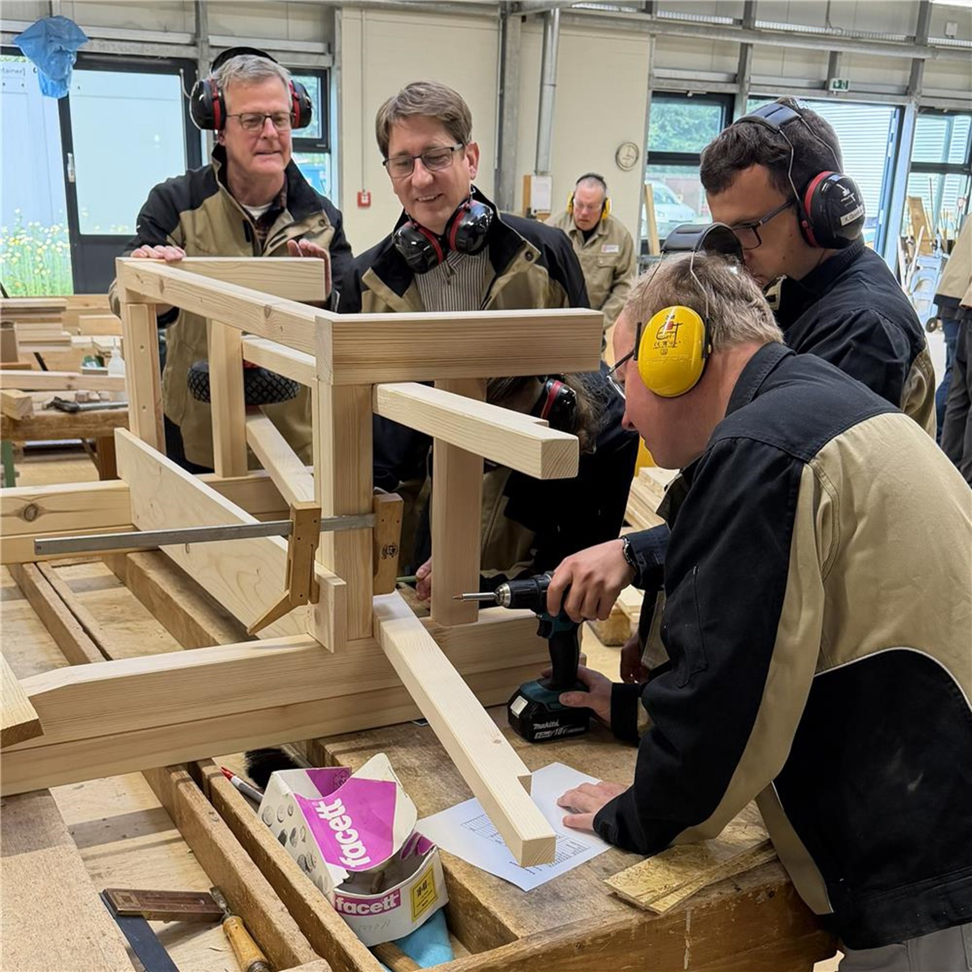 Mehrere Personen arbeiten gemeinsam in einer Werkstatt an einer Holzkonstruktion. Mit Werkzeugen und Schutzausrüstung wird das Holz bearbeitet, während die Gruppe den Arbeitsschritt abstimmt.