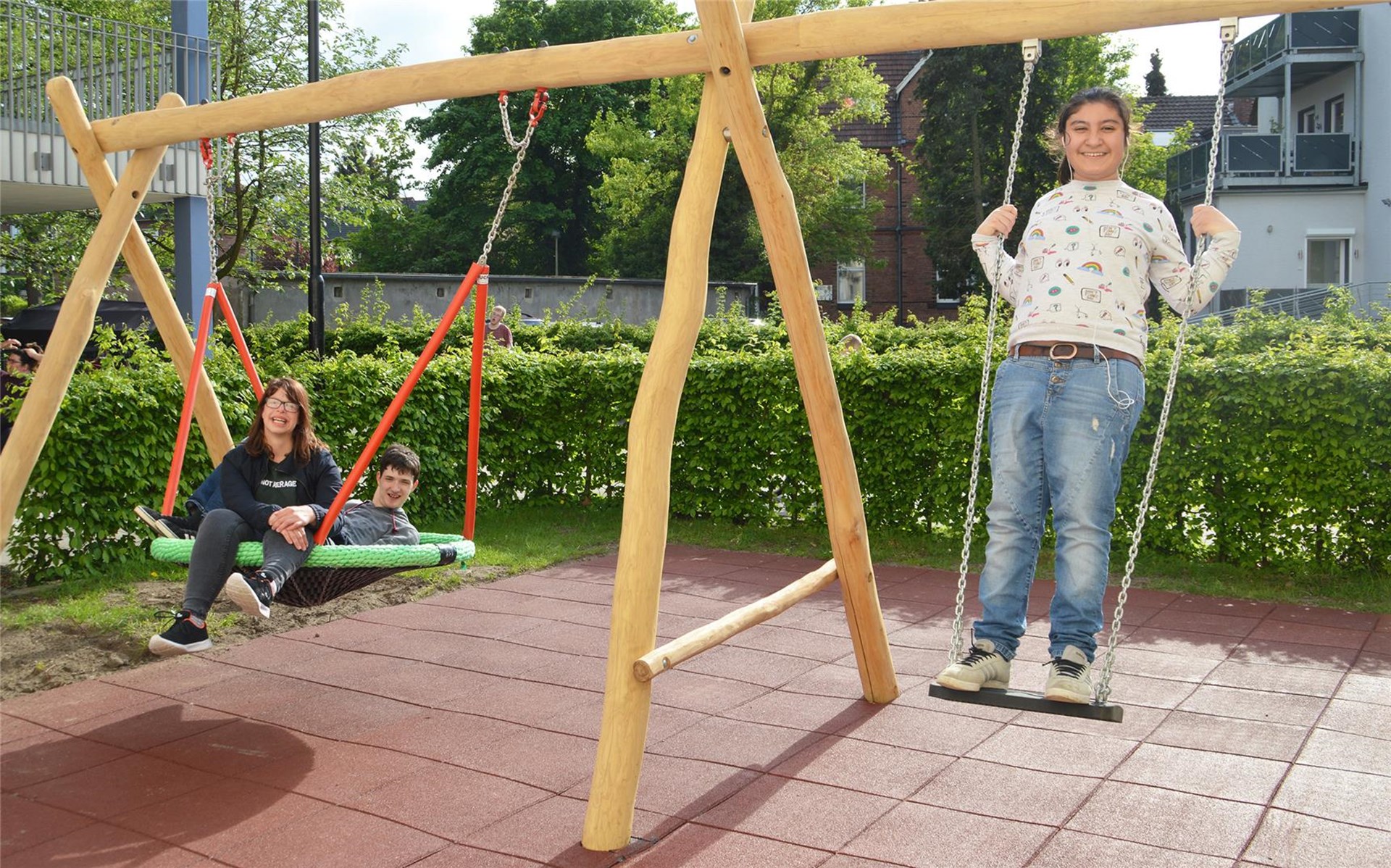 Zwei Kinder sitzen in einer Nestschaukel, ein weiteres Kind steht auf einer normalen Schaukel