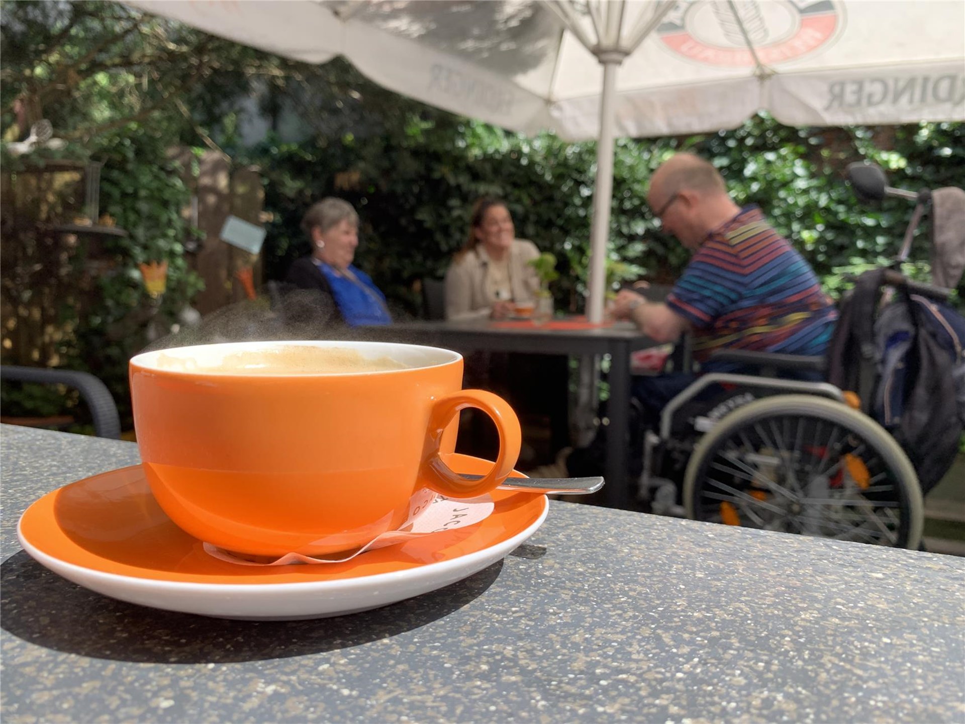 Eine Tasse mit Untertasse auf einem Tisch. Im Hintergrund ist ein Tisch mit Sonnenschirm und drei Personen zu sehen, von denen einer im Rollstuhl sitzt