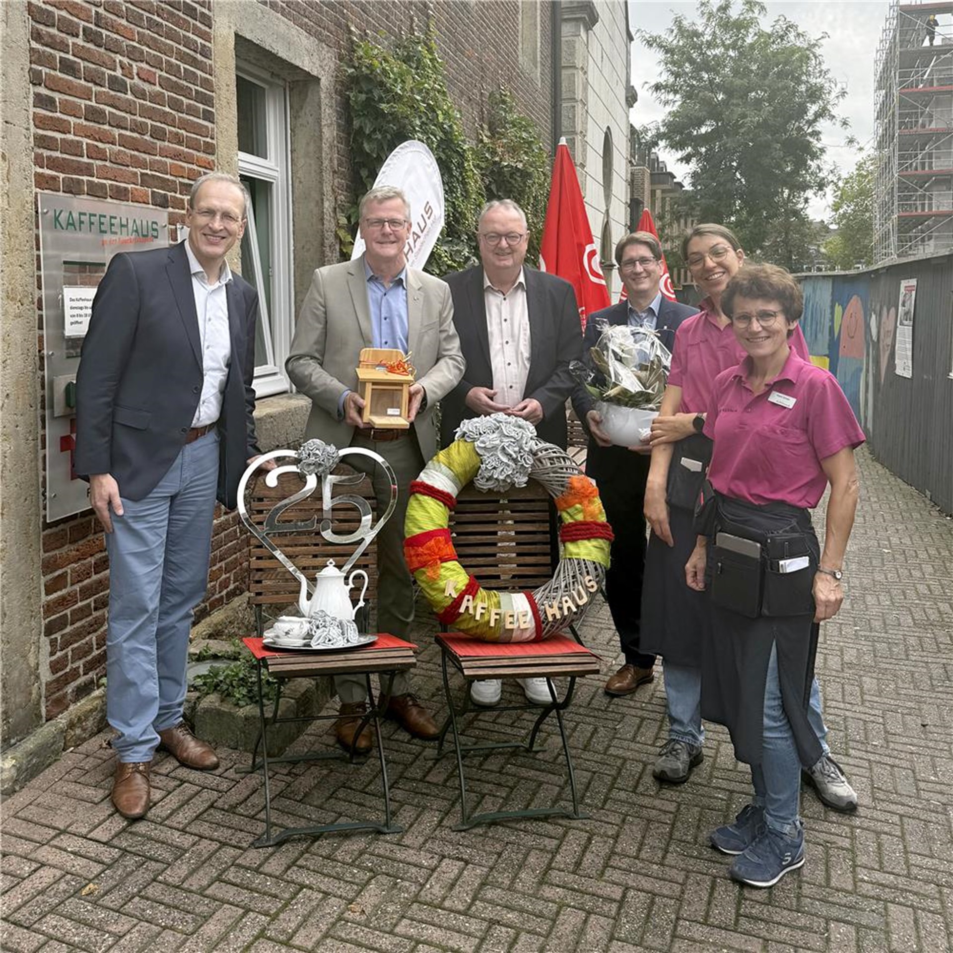 Sechs Personen stehen vor dem Eingang des Kaffeehauses für ein Gruppenfoto. Vor ihnen sind eine Dekoration zum 25-jährigen Jubiläum, ein großer Kranz sowie Blumen und Geschenke aufgestellt.