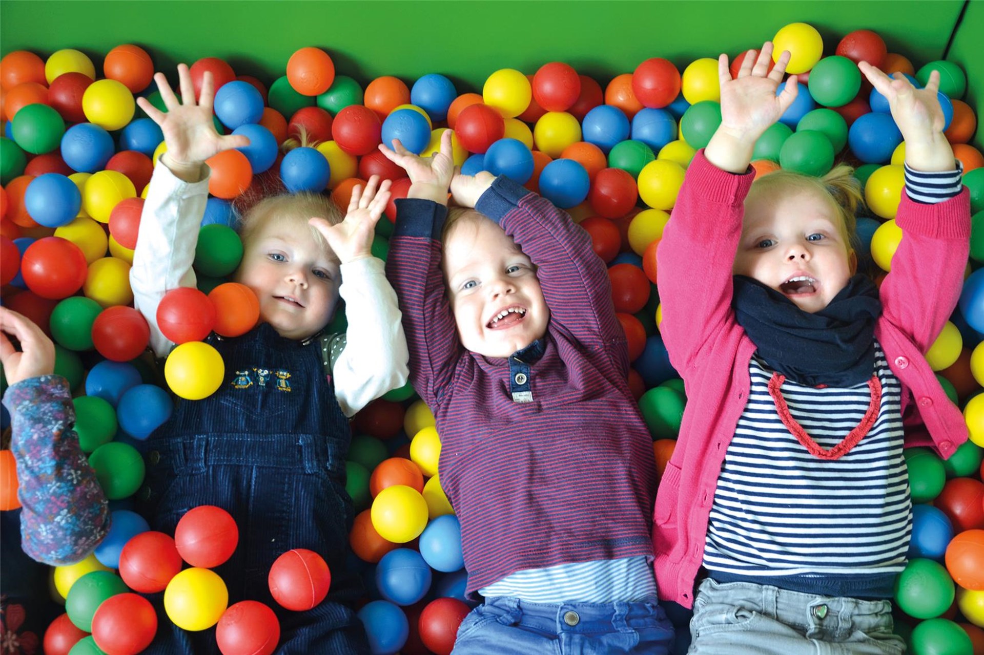 Drei Kinder liegen auf dem Rücken im Bällebad und strecken die Hände nach oben.