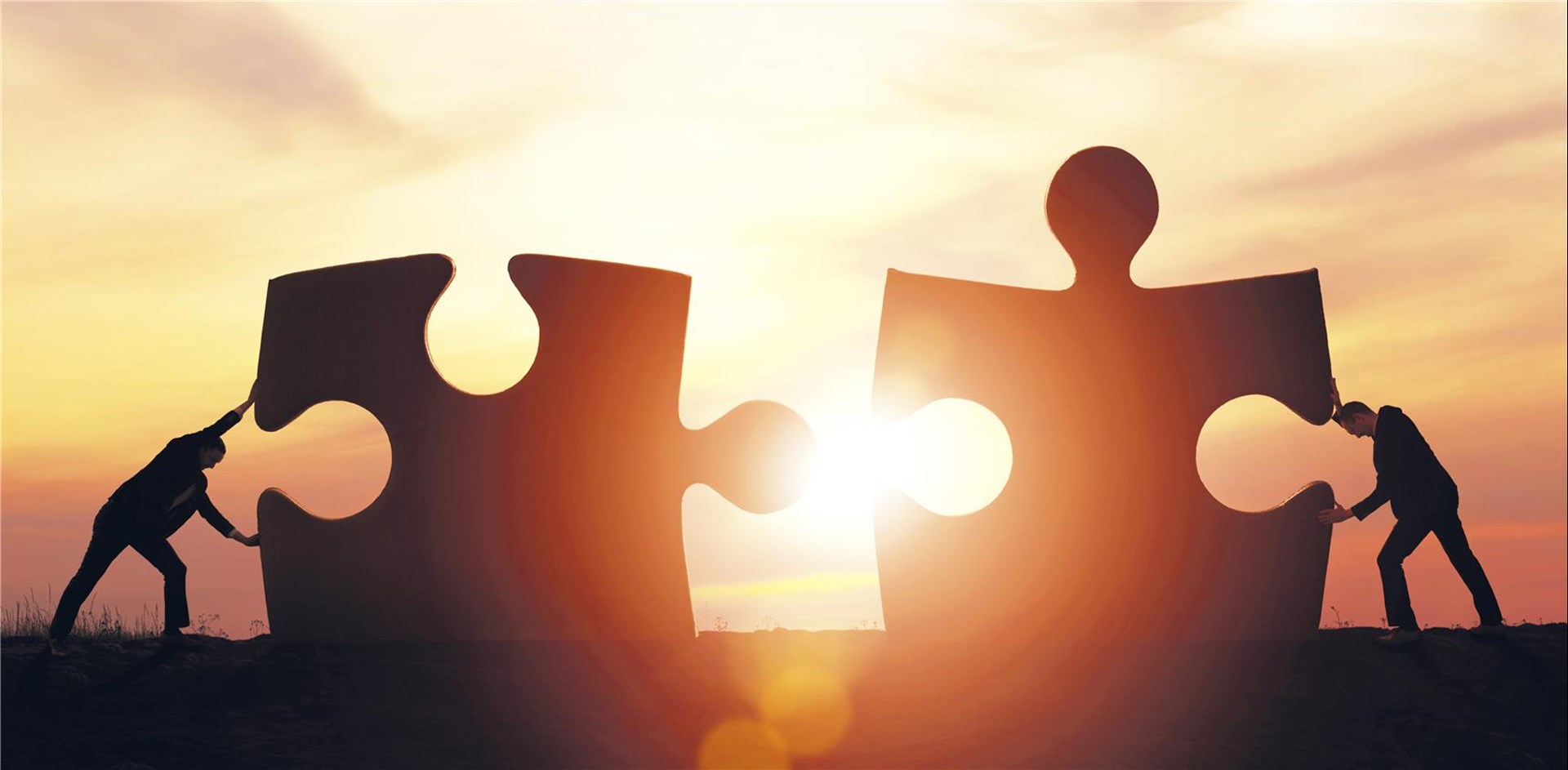 Zwei sehr große Puzzleteile, dir vor einem Sonnenuntergang von zwei Personen zusammengeschoben werden.