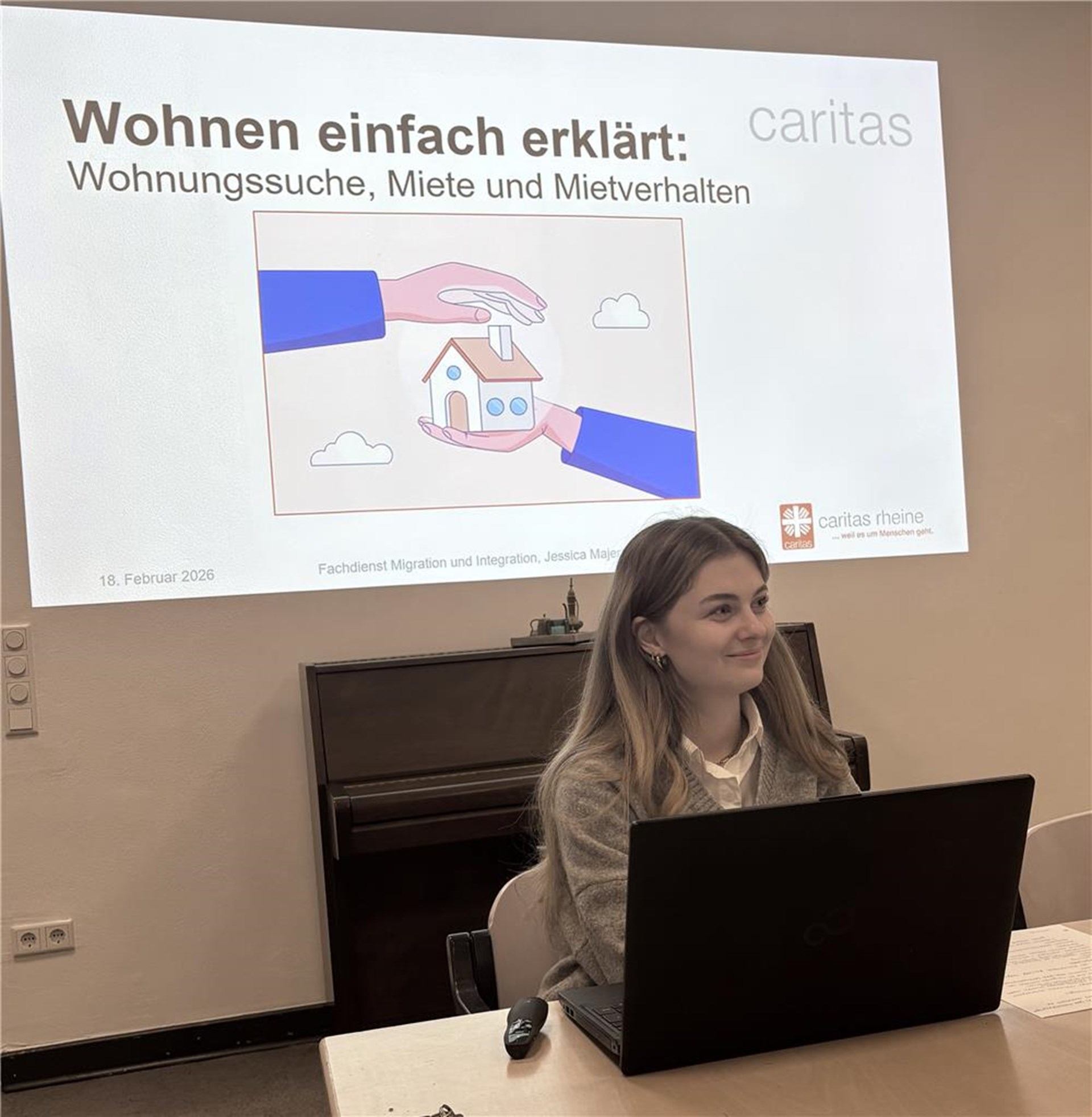 Eine Frau sitzt an einem Tisch vor einem aufgeklappten Laptop und hält eine Präsentation. Auf der Leinwand hinter ihr ist eine Folie mit dem Titel „Wohnen einfach erklärt: Wohnungssuche, Miete und Mietverhalten“ zu sehen. 