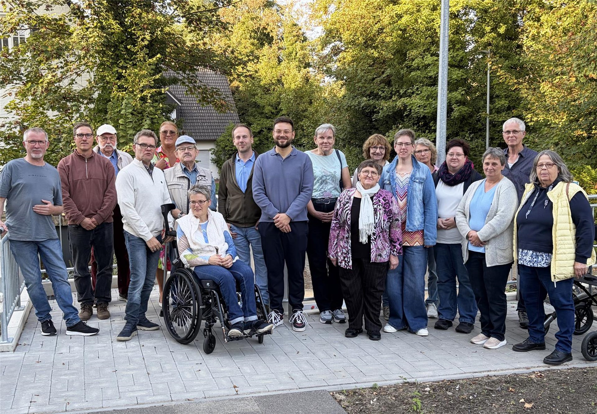 Gruppenfoto von rund 18 Personen auf einem gepflasterten Platz im Freien. Die Gruppe steht und sitzt, eine Person im Rollstuhl, umgeben von Bäumen und herbstlichem Grün.