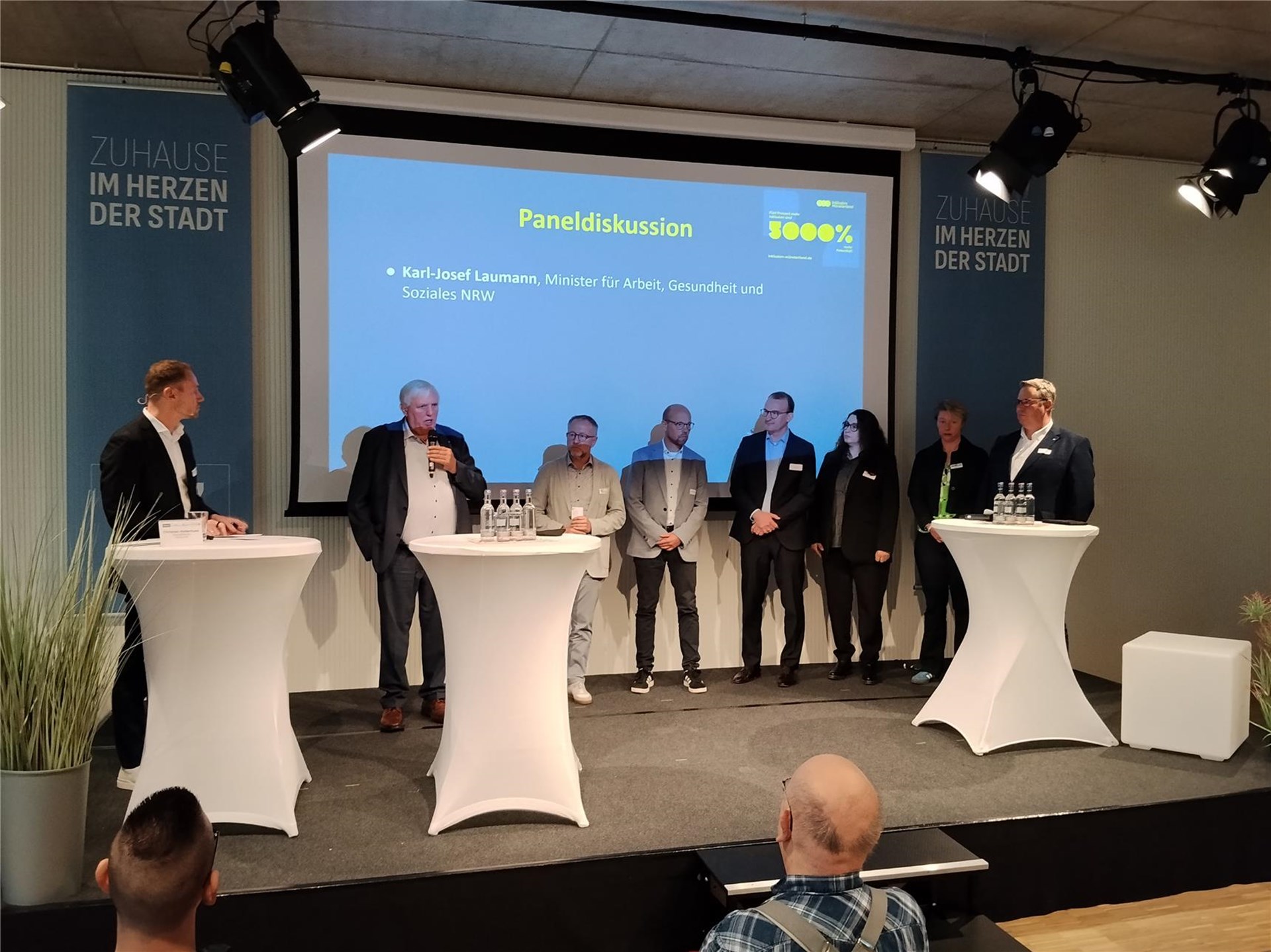 Mehrere Personen stehen auf einer Bühne bei einer Podiumsdiskussion. Im Hintergrund zeigt eine große Leinwand die Veranstaltung, vor den Teilnehmenden stehen Stehtische mit Mikrofonen und Getränken.