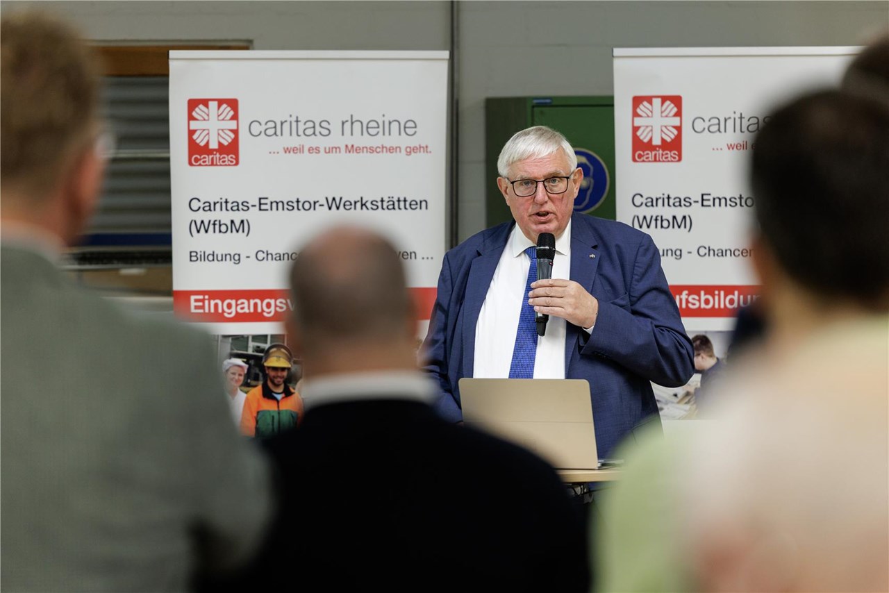 Karl-Josef Laumann hält vor Zuhörerinnen und Zuhörern einen Vortrag mit Mikrofon. Im Hintergrund sind Roll-up-Banner der Caritas-Emstor-Werkstätten zu sehen.